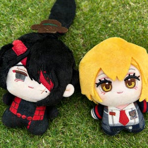 Peut inclure: Deux poupées en peluche de style anime. L'une a des cheveux noirs avec des détails rouges et un petit chapeau. L'autre a des cheveux jaunes et porte un uniforme scolaire avec une cravate rouge. Les deux poupées ont de grands yeux et sont sur une surface herbeuse verte.