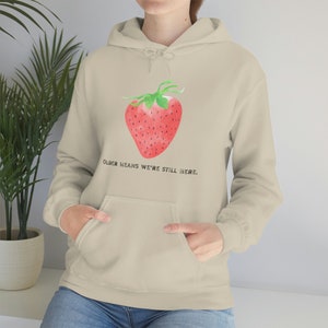 Könnte beinhalten: Ein hellbeiger Hoodie mit einem roten und grünen Erdbeer-Motiv und dem Text "Older means we're still here."