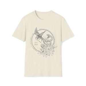 May include: Cream-colored t-shirt featuring a detailed black graphic of a bird attacking a snake, surrounded by floral elements. The design includes the text "nothing you can take from me was ever worth keeping" and the date "11.17 2025".