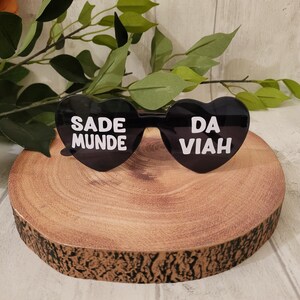 May include: Black heart-shaped sunglasses with white text that reads "SADE MUNDE" on the left lens and "DA VIAH" on the right lens. The sunglasses are displayed on a round, wooden surface with a natural edge, with green leaves in the background.