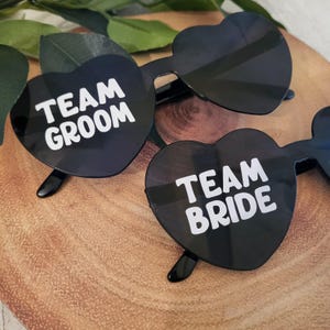 May include: Two pairs of black heart-shaped sunglasses with white text. One pair says "Team Groom" and the other says "Team Bride".