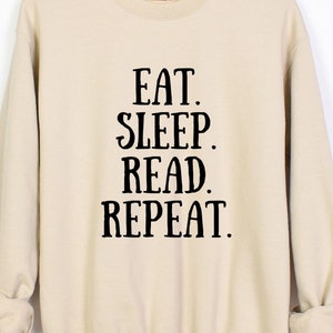 May include: A beige sweatshirt with the words "EAT. SLEEP. READ. REPEAT." printed in black. The sweatshirt is hanging on a wooden hanger against a white background. The sleeves are cuffed.
