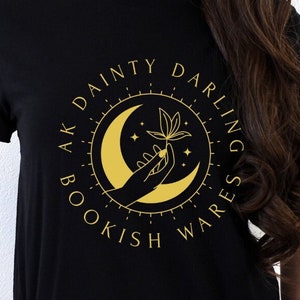 Puede incluir: Camiseta negra con un diseño circular dorado que presenta una luna creciente, una mano y elementos botánicos. El texto "AK DAINTY DARLING BOOKISH WARES" está impreso alrededor del diseño. La camiseta la lleva una persona.