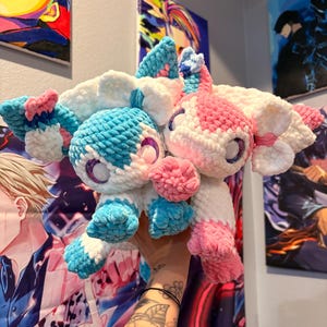 May include: Two handmade plush toys, one blue and white, the other pink and white. Both have large, fluffy ears and large, glittery eyes. The toys are held up in front of a wall with colorful artwork.