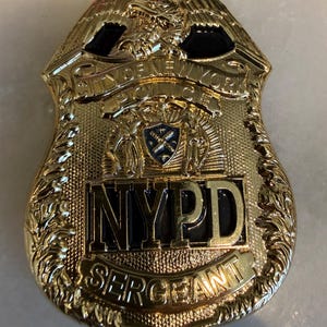 May include: A gold-toned metal NYPD Sergeant badge featuring an eagle at the top, the text CITY OF NEW YORK POLICE, a central shield emblem with blue accents, the letters NYPD in a prominent black rectangular field, and the word SERGEANT below.
