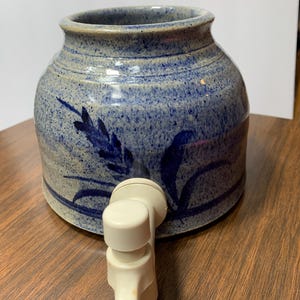 May include: A ceramic water dispenser with a blue and beige speckled finish. The dispenser features a floral design and a white spigot. The stoneware container is a round shape with a wide opening.