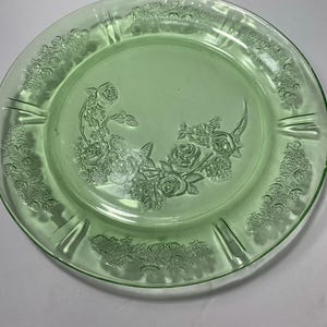 May include: A round, light green glass plate featuring a pressed floral pattern. The center displays a cluster of embossed roses and leaves, while the outer rim is decorated with a repeating pattern of small flowers separated by smooth, vertical indentations.