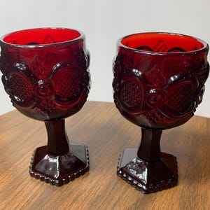 May include: Two ruby red goblets with ornate, embossed designs. The goblets have a detailed pattern on the bowl and a square base. The stem and base are a deep red, and the interior of the bowl is a bright red.