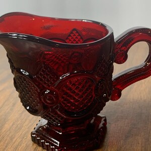 May include: A ruby red glass creamer with ornate, textured detailing. The creamer features a handle and a square base. The design includes diamond and floral patterns. The creamer is likely vintage or antique.