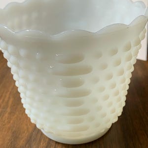 May include: A white milk glass bowl featuring a hobnail pattern across the exterior surface. The bowl has a scalloped rim and a rounded base, with a translucent quality to the glass. The texture consists of raised, rounded bumps in rows.