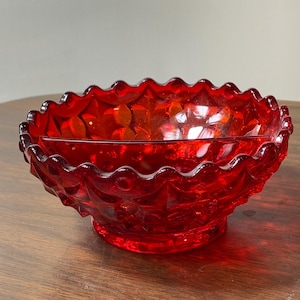 May include: A ruby red glass bowl with a scalloped rim and a decorative pattern. The bowl has a round base and a textured surface. The bowl is likely vintage and suitable for serving or display.