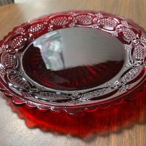 May include: A ruby red glass plate with a scalloped edge and intricate embossed detailing. The plate has a central, smooth surface surrounded by a decorative border. The plate is likely vintage or antique.