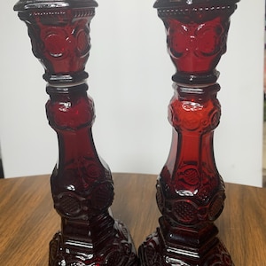 May include: Two matching ruby red glass candle holders. Each holder has a square base, a detailed stem, and a flared top. The glass has an embossed floral and scroll design. The candle holders are displayed on a wooden surface.