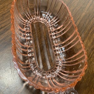 May include: A pink glass serving dish with an oval shape and ribbed sides. The dish has decorative handles and a scalloped edge. The glass is transparent, allowing the wooden surface beneath to be partially visible.