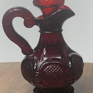 May include: A ruby red glass syrup pitcher with a decorative handle and a textured, mushroom-shaped stopper. The pitcher features embossed floral and geometric patterns. The base is square and also has a decorative pattern.