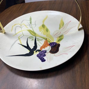 May include: Oval ceramic serving dish with a gold handle. The dish features a hand-painted design of stylized fruit and foliage in shades of green, yellow, orange, purple, and black. A decorative piece for serving snacks or displaying fruit.