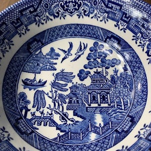May include: A blue and white ceramic plate with a Willow pattern design. The plate features a detailed landscape scene with pagodas, bridges, and birds. The rim has a decorative border. The plate is a classic piece of tableware.