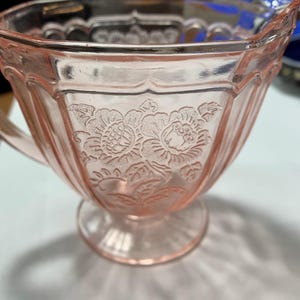 May include: A pink glass creamer with a floral embossed design. The creamer has a handle and a pedestal base. The glass is a soft pink color and has a vintage look. The creamer is likely part of a vintage glassware set.