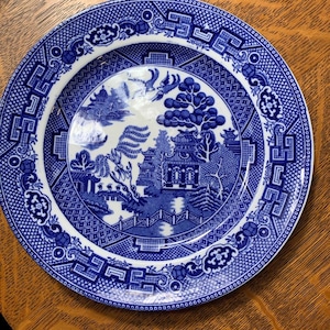 May include: A blue and white ceramic plate with a Willow pattern design. The plate features a detailed landscape scene with a bridge, buildings, and trees. The rim of the plate has a decorative border. The plate is set on a wooden surface.