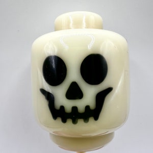 May include: A light-colored, plastic skull head with black eyes and a grinning mouth. The head is a Halloween-themed decoration, likely for a toy or craft project. The head is a light cream color.