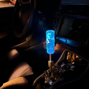 Puede incluir: Una palanca de cambios con un patr&oacute;n azul y blanco, con una carcasa de pl&aacute;stico transparente y una base de metal plateado. La palanca est&aacute; iluminada desde el interior, creando un efecto brillante.