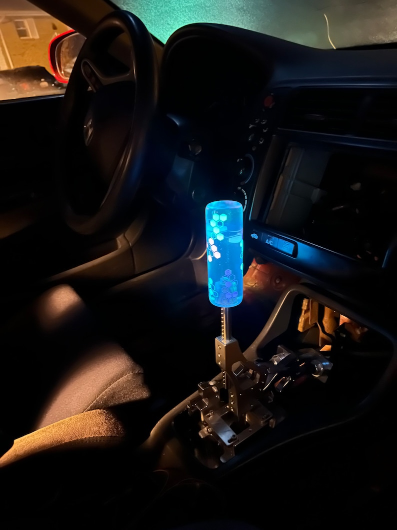 Puede incluir: Una palanca de cambios con un patr&oacute;n azul y blanco, con una carcasa de pl&aacute;stico transparente y una base de metal plateado. La palanca est&aacute; iluminada desde el interior, creando un efecto brillante.