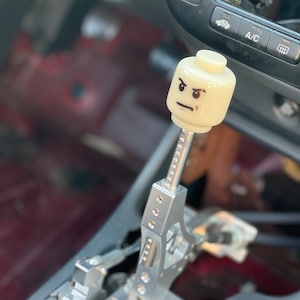 Op de afbeelding: Een zilveren versnellingspook met een wit Lego minifiguur hoofd erop. Het minifiguur heeft een zwarte boze uitdrukking.