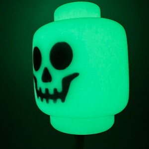May include: A glowing green Lego-style head with a black skull design. The Halloween-themed decoration has large eye sockets and a detailed jawline. The head is a light green color and glows in the dark.