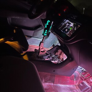 Puede incluir: Una vista cercana del interior de un coche con una palanca de cambios personalizada. La palanca es negra con un patr&oacute;n verde que brilla en la oscuridad. El interior del coche es rojo y negro.