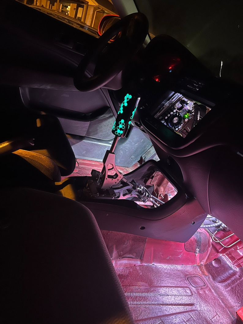 Puede incluir: Una vista cercana del interior de un coche con una palanca de cambios personalizada. La palanca es negra con un patr&oacute;n verde que brilla en la oscuridad. El interior del coche es rojo y negro.