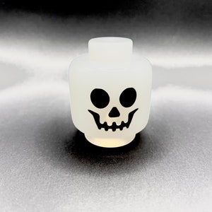 May include: A white plastic skull head with black eyes and a grinning mouth. The head is a toy or decorative item, likely for Halloween or themed events. The design is simple and bold, with a cartoonish aesthetic.