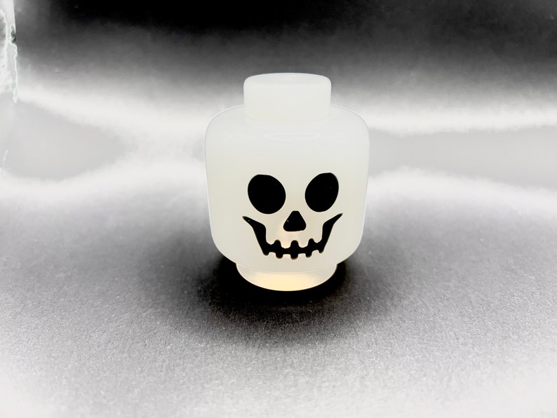 May include: A white plastic skull head with black eyes and a grinning mouth. The head is a toy or decorative item, likely for Halloween or themed events. The design is simple and bold, with a cartoonish aesthetic.