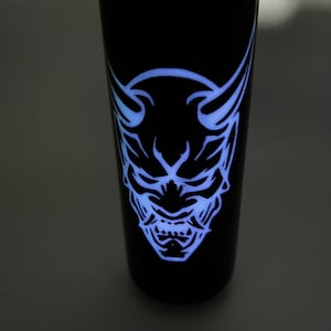 Könnte beinhalten: Schwarzer Becher mit einem leuchtend blauen Oni-Masken-Design. Die Maske hat spitze Hörner, einen strengen Ausdruck und komplizierte Details. Das Design leuchtet hell vor dem dunklen Hintergrund und erzeugt einen auffälligen visuellen Kontrast.