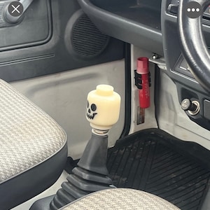 May include: A custom gear shift knob in a vehicle interior. The knob is a light yellow Lego brick with a black skull face. The shift boot is black, and the interior has gray and patterned fabric.