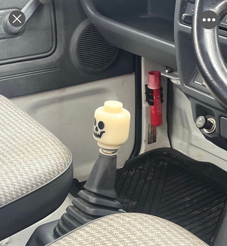 May include: A custom gear shift knob in a vehicle interior. The knob is a light yellow Lego brick with a black skull face. The shift boot is black, and the interior has gray and patterned fabric.