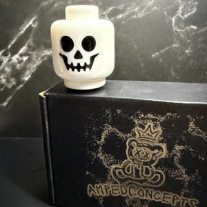 May include: A white Lego-style head with a black skull face. The head is sitting on a black box with a gold bear logo and the text "AMPEDCONCEPTS". The background is a dark gray marble pattern.