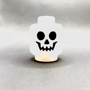 May include: A white skull-shaped decorative object with black eyes and a mouth. The object is a stylized representation of a skull, perfect for Halloween or spooky-themed decor. The object is illuminated from within, creating a soft glow.