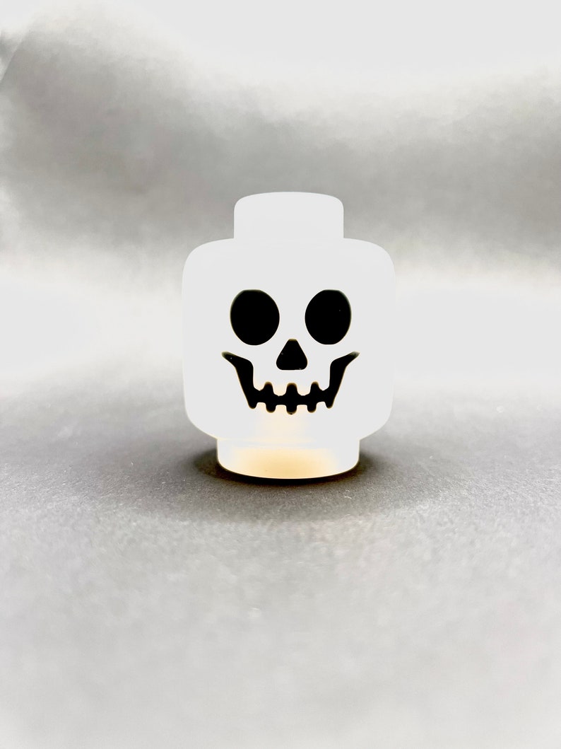 May include: A white skull-shaped decorative object with black eyes and a mouth. The object is a stylized representation of a skull, perfect for Halloween or spooky-themed decor. The object is illuminated from within, creating a soft glow.