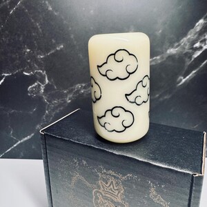 K&ouml;nnte beinhalten: Eine wei&szlig;e Kerze mit schwarzen Wolkenmotiven. Die Kerze steht auf einer schwarzen Box mit einem Logo.