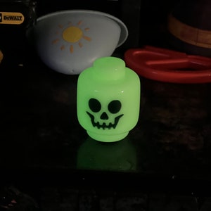 May include: A glowing green Lego-style head with a black skull face. The toy is a novelty item, likely for Halloween or spooky-themed decor. The head is the primary focus, with other objects in the background.