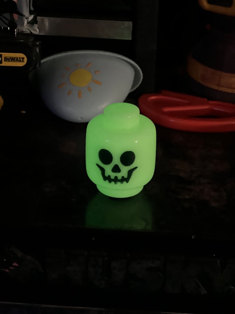 May include: A glowing green Lego-style head with a black skull face. The toy is a novelty item, likely for Halloween or spooky-themed decor. The head is the primary focus, with other objects in the background.
