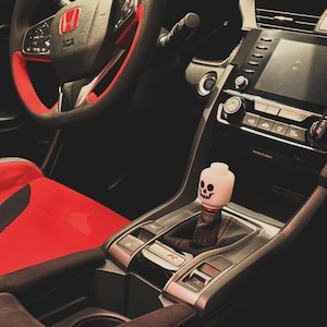 Op de afbeelding: Interieur van een Honda Civic Type R. Het stuurwiel is zwart met rode accenten en de stoelen zijn rood. Een aangepaste versnellingspookknop met een schedelontwerp is zichtbaar. Het dashboard is zwart met een digitaal scherm.