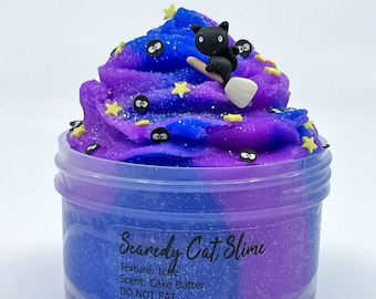 May include: A jar of "Scaredy Cat Slime" with a blue and purple color scheme. The slime is decorated with a black cat riding a broom, yellow stars, and black eye-shaped sprinkles. The text on the jar reads "Texture: Icee, Scent: Cake Batter, DO NOT EAT."