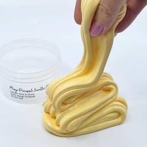 May include: A yellow slime with a glossy finish, being stretched and pulled from a clear plastic container. The slime is labelled "Mango Pineapple Smoothie &" with the text "Texture: Thick & Glossy, Scent: Mango Pineapple, DO NOT EAT".