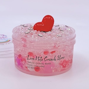 May include: A clear plastic jar filled with pink slime, decorated with a red heart and small, silver shapes. The jar is labelled "Love Note Crunch Slime." The texture is "Crunchy Bomb" and the scent is "Unscented." The label also says "DO NOT EAT."