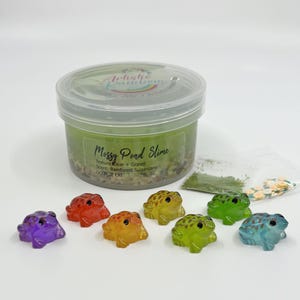 May include: A clear plastic container of "Messy Pond Slime" with a green hue, gravel, and a variety of colorful frog figurines. The slime is labeled with the texture, scent, and a warning to not eat it.