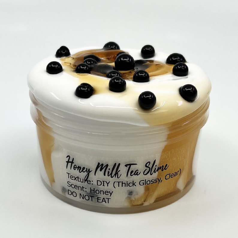 May include: A clear plastic container filled with white and brown slime with black boba pearls on top. The container has a label that reads "Honey Milk Tea Slime", "Texture: DIY (Thick Glossy, Clear)", "Scent: Honey", and "DO NOT EAT".