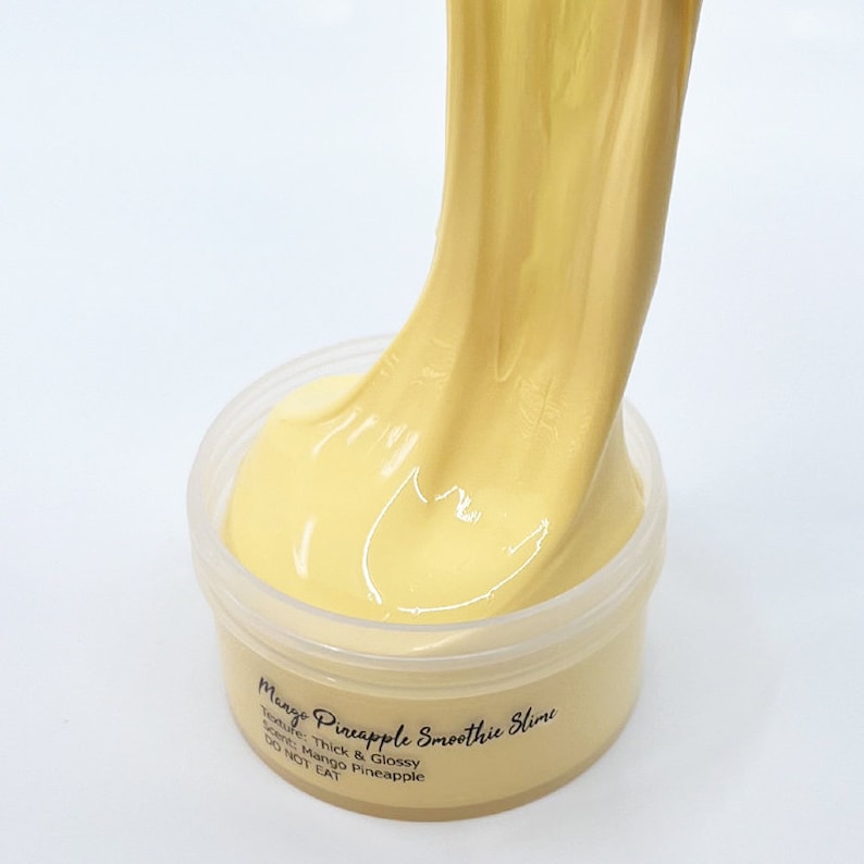 May include: A close-up of a yellow slime in a clear plastic container. The slime is being pulled out of the container and is thick and glossy. The label on the container reads "Mango Pineapple Smoothie Slime", "Texture: Thick & Glossy", "Scent: Mango Pineapple", and "DO NOT EAT".