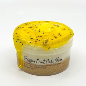 May include: A clear plastic jar filled with Passion Fruit Cake Slime. The slime has a yellow, glossy top layer with black specks, resembling passion fruit seeds, dripping down the sides. The jar has a white and brown layered appearance with text that reads "Passion Fruit Cake Slime".