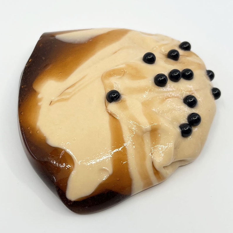 Puede incluir: Primer plano de un slime marr&oacute;n claro y beige con remolinos marrones oscuros y cuentas negras. El slime est&aacute; sobre una superficie blanca. El slime es un juguete.